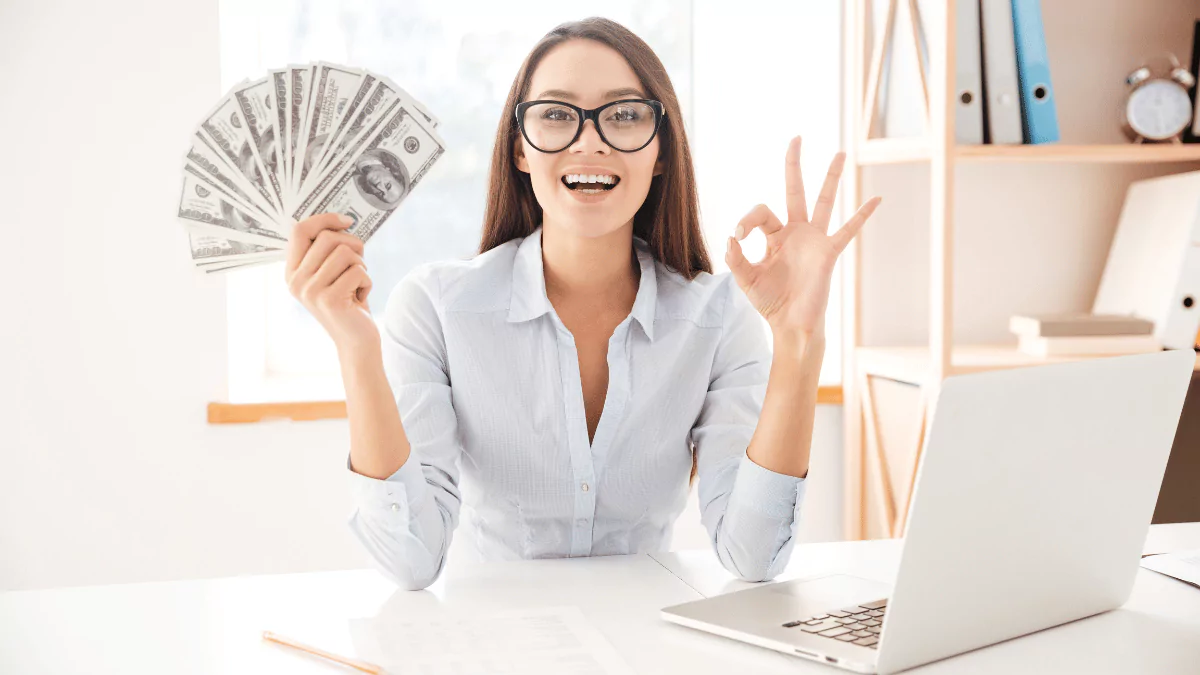 Businesswoman in a white shirt sitting in her office, holding cash and making an OK gesture