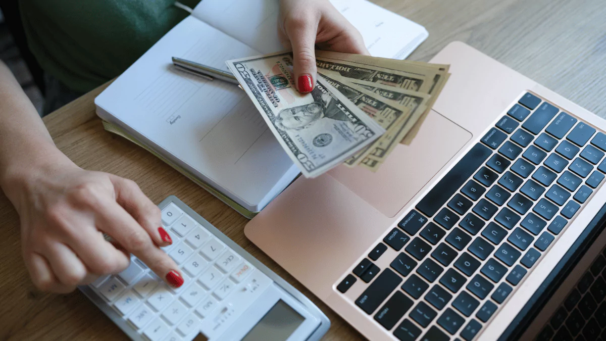 hands are holding money while using a laptop and a calculator