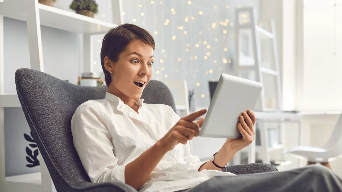 Overjoyed Adult Woman Reading Exciting News on Tablet Sitting in Comfortable Armchair at Home